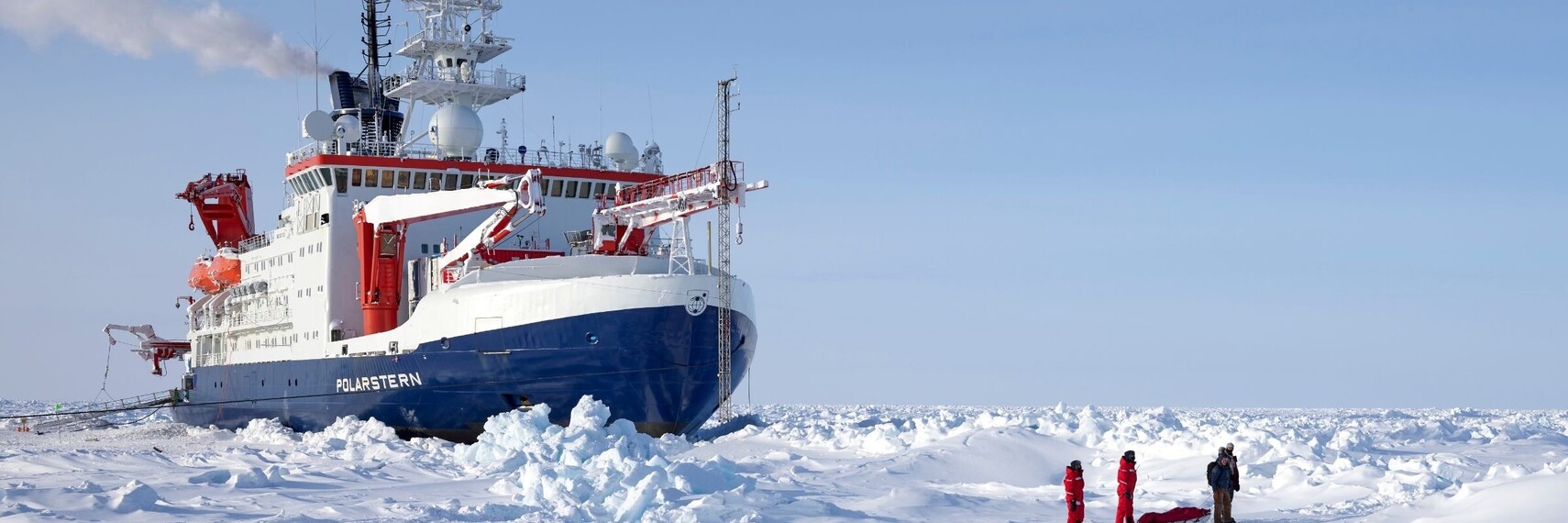 Der Eisbrecher Polarstern im arktischen Eis und drei Wissenschaftler stehen in Eisfeld davor.
